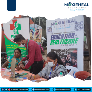 Volunteer in a red shirt assists a seated woman at a clinic table during a Moxieheal Foundation outreach event with health banners behind them.
