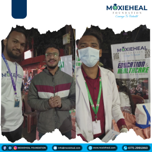 Two men at a Moxieheal Foundation event posing for a photo; one in a gray jacket, the other in a white coat with a mask, banners in the background.