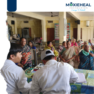 Two doctors in white coats sit at a table with medical equipment while elderly people wait in a hall, a health camp scene.",